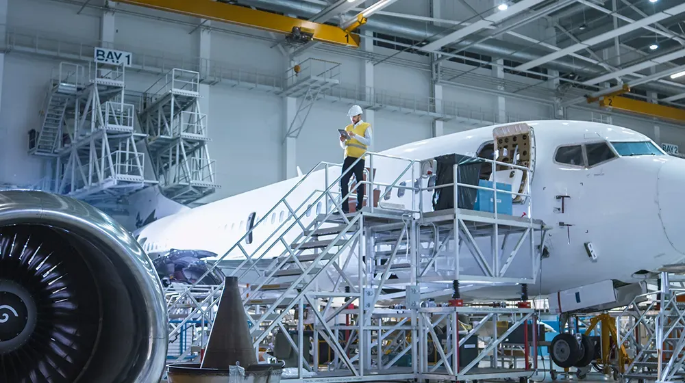 A person in a hard hat and safety vest inspects an airplane fuselage in a manufacturing hangar, using a tablet. The scene highlights modern aircraft production and maintenance within a B2B manufacturing environment.