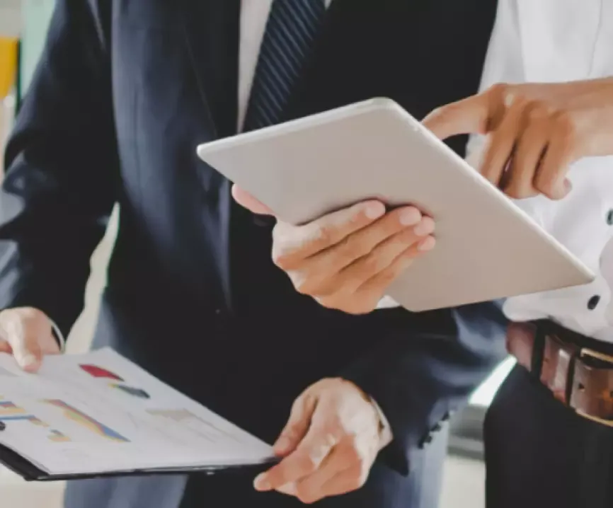 Two business professionals in suits review a digital tablet and financial charts. One points to the tablet screen, indicating collaboration and data analysis for manufacturing operations in a modern office setting.