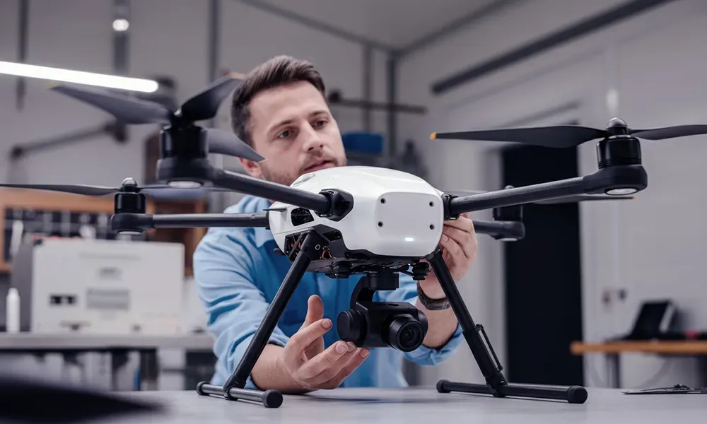 Man in a blue shirt inspects a white drone with black propellers and a camera gimbal in a workshop setting. Manufacturing technology.