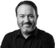 Black and white headshot of Zane Shewalter, smiling and looking at the camera. He has a beard and is wearing a black collared shirt.