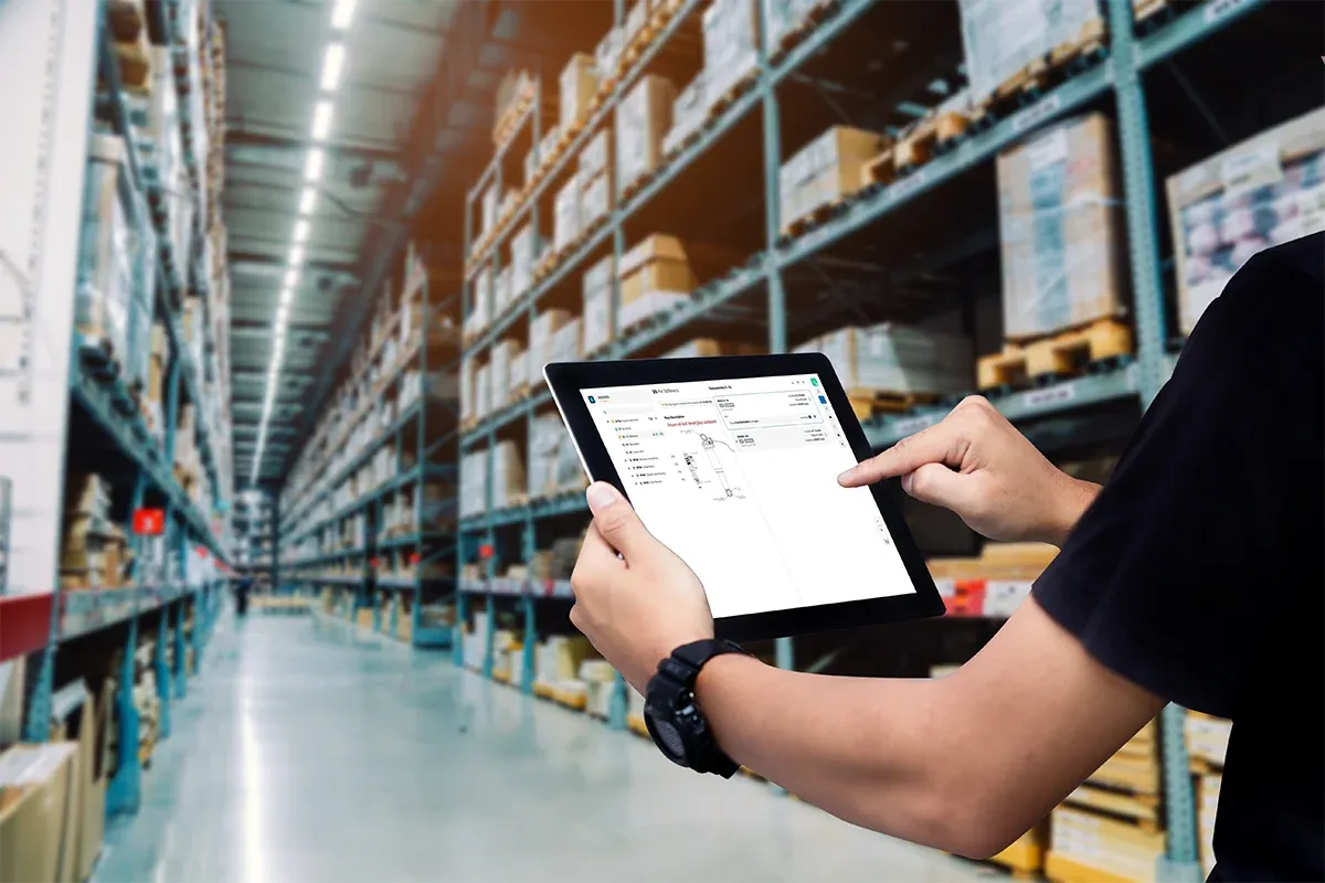 Close-up of a person's hands holding a tablet displaying an interface with product information. The background is a blurred modern warehouse with tall shelves stacked with boxes.