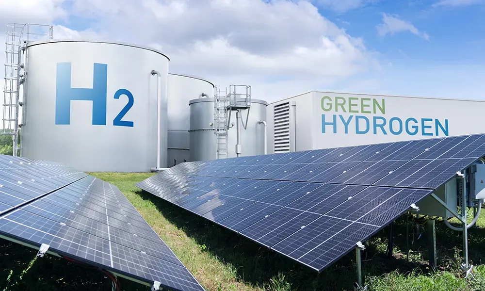 Industrial plant with large H2 storage tanks and a green hydrogen production building, in front of a field of solar panels under a cloudy blue sky.