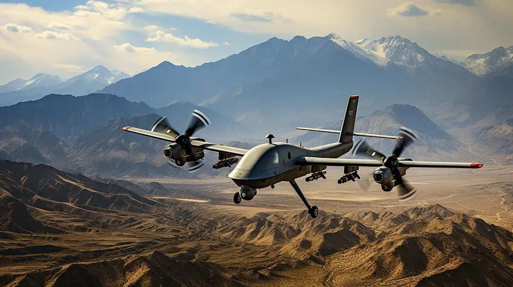 A grey military drone with two propellers flies over a dry, mountainous landscape with snow-capped mountains in the background under a cloudy sky. The drone carries munitions.