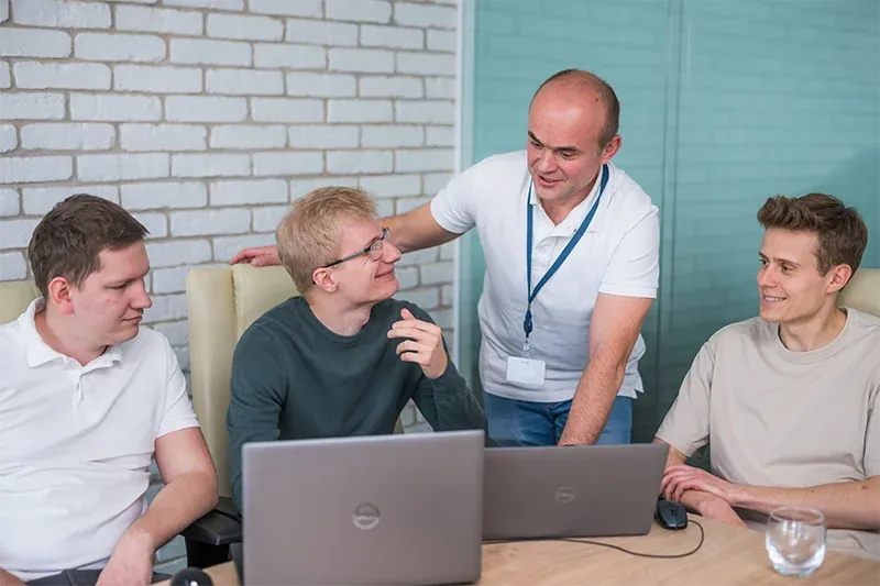 Four men collaborate around two laptops on a wooden table, in an office with a white brick wall and blue glass partition. One man in a white polo shirt leans over, talking to a man in glasses, who smiles. Two other men watch on either side.