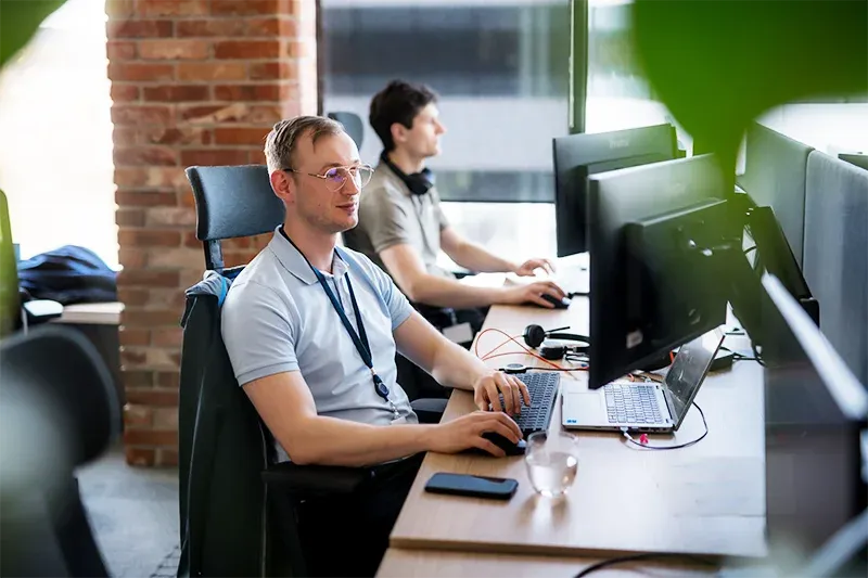 Two men in a modern office setting work at computers. One man in a light blue polo shirt and glasses types on a keyboard, looking focused. Another man in a t-shirt and headphones works in the background. The scene suggests productivity and a collaborative work environment.