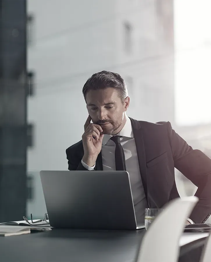 A focused businessman in a suit and tie studies a laptop on a desk in a modern office. He rests his chin on his hand, contemplating. Includes a glass of water and other office items on the desk.